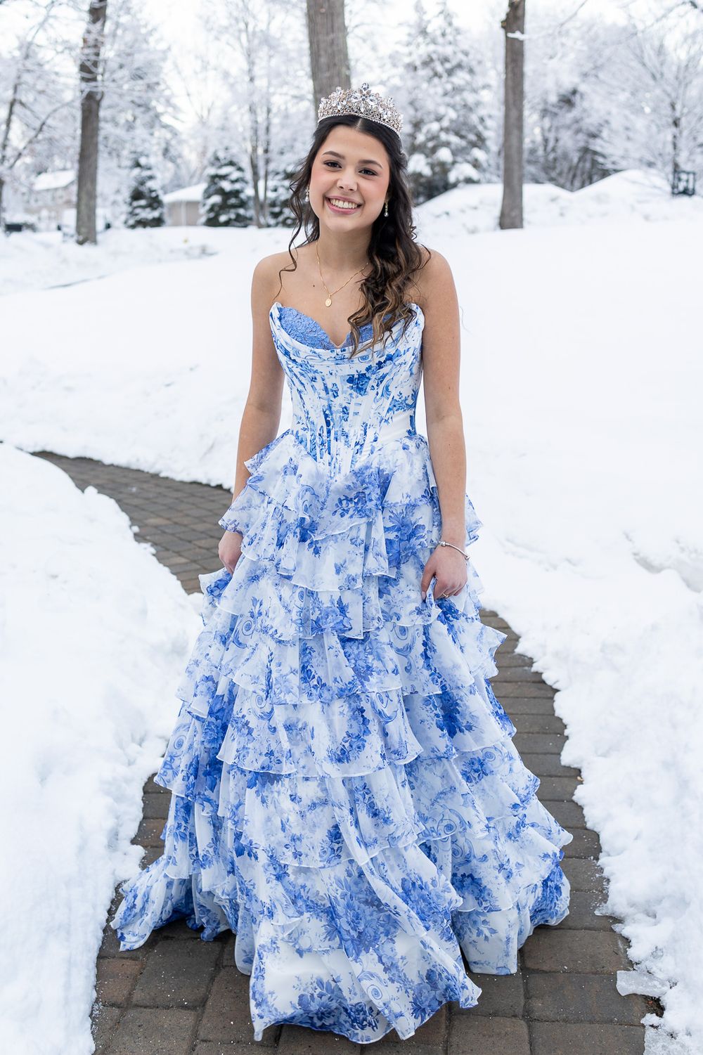 Young woman in blue floral ball gown and tiara smiling on snowy brick path outdoors.
