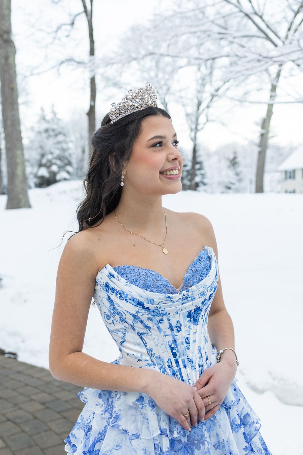 Young woman in blue floral ballgown and tiara smiles outdoors in snowy winter setting.