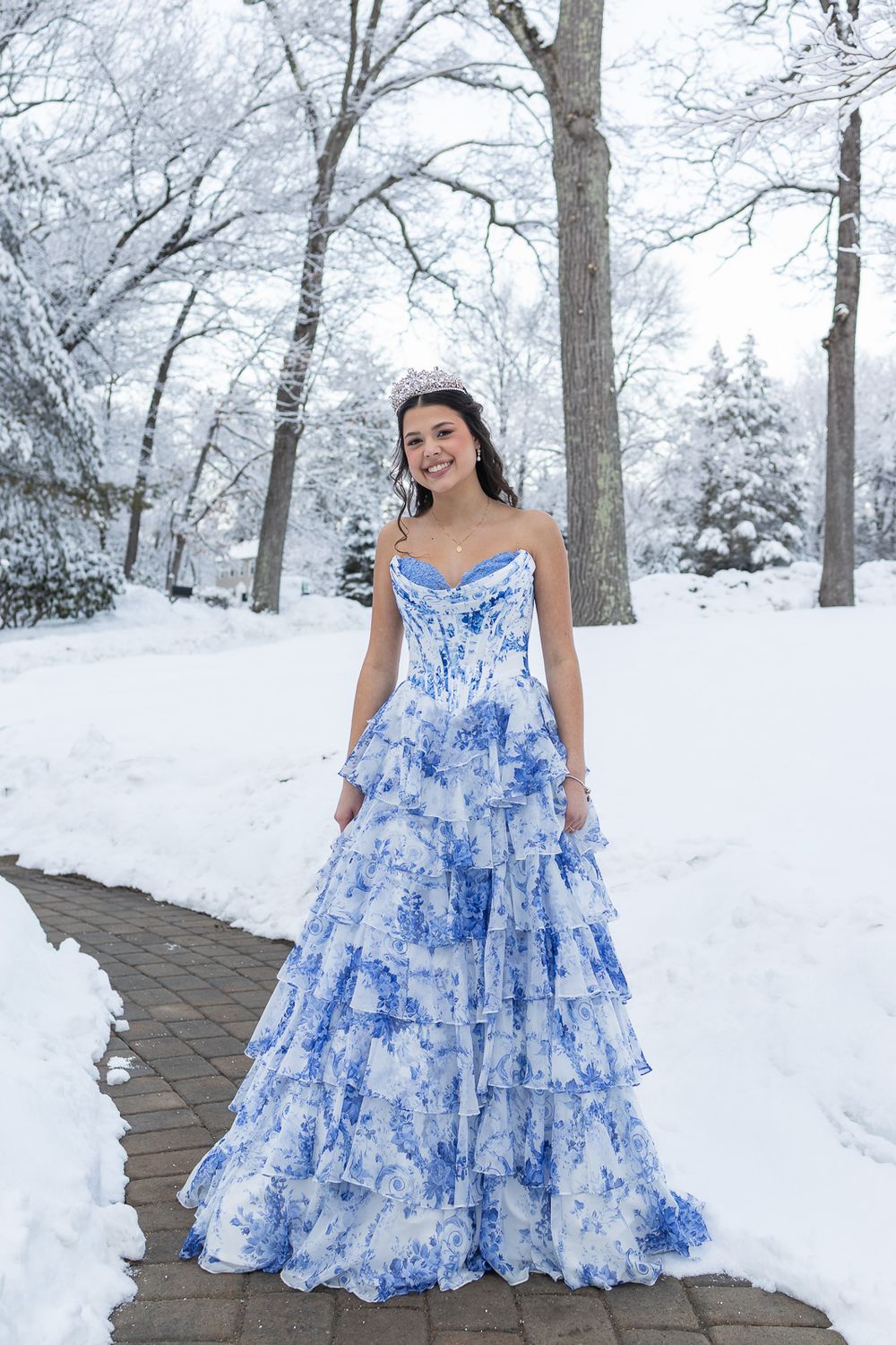 Young woman in a blue floral strapless ball gown posing on a snowy winter path with snow-covered trees.