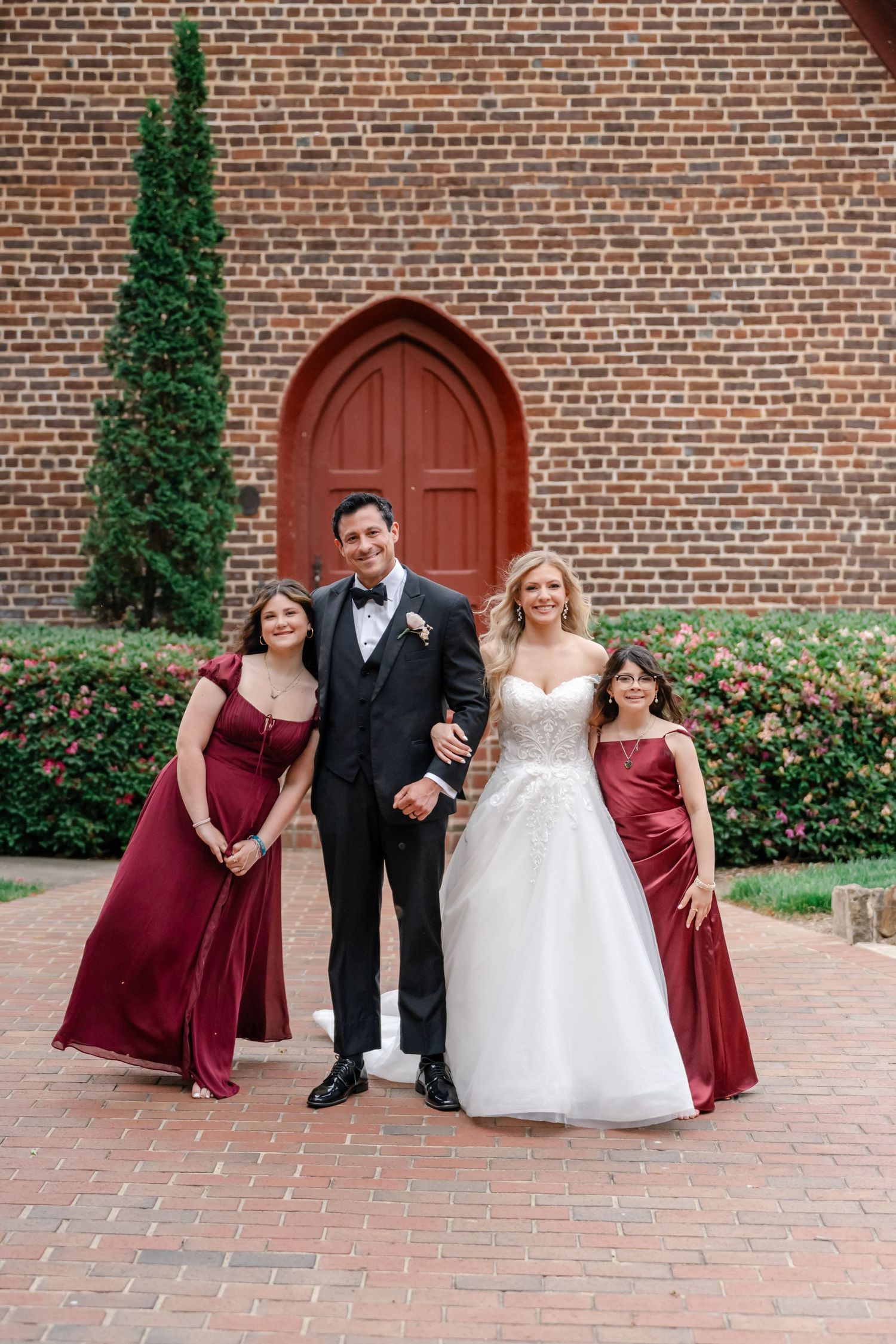 Bride in white gown and groom in black tux pose with two bridesmaids in burgundy dresses outside brick church.