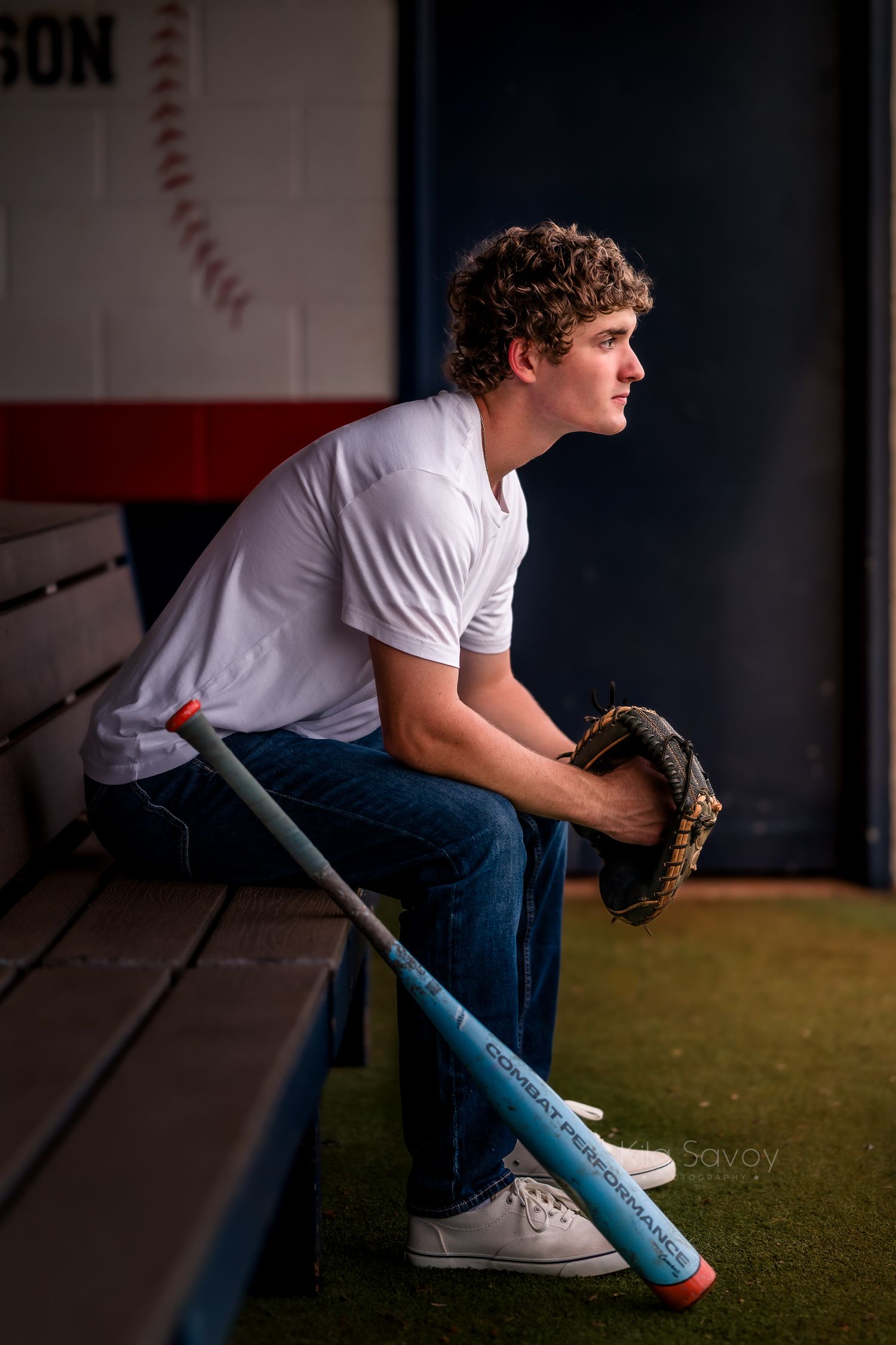 Young baseball player sits in dugout holding glove and bat, wearing white tee and jeans, gazing thoughtfully into the distance.
