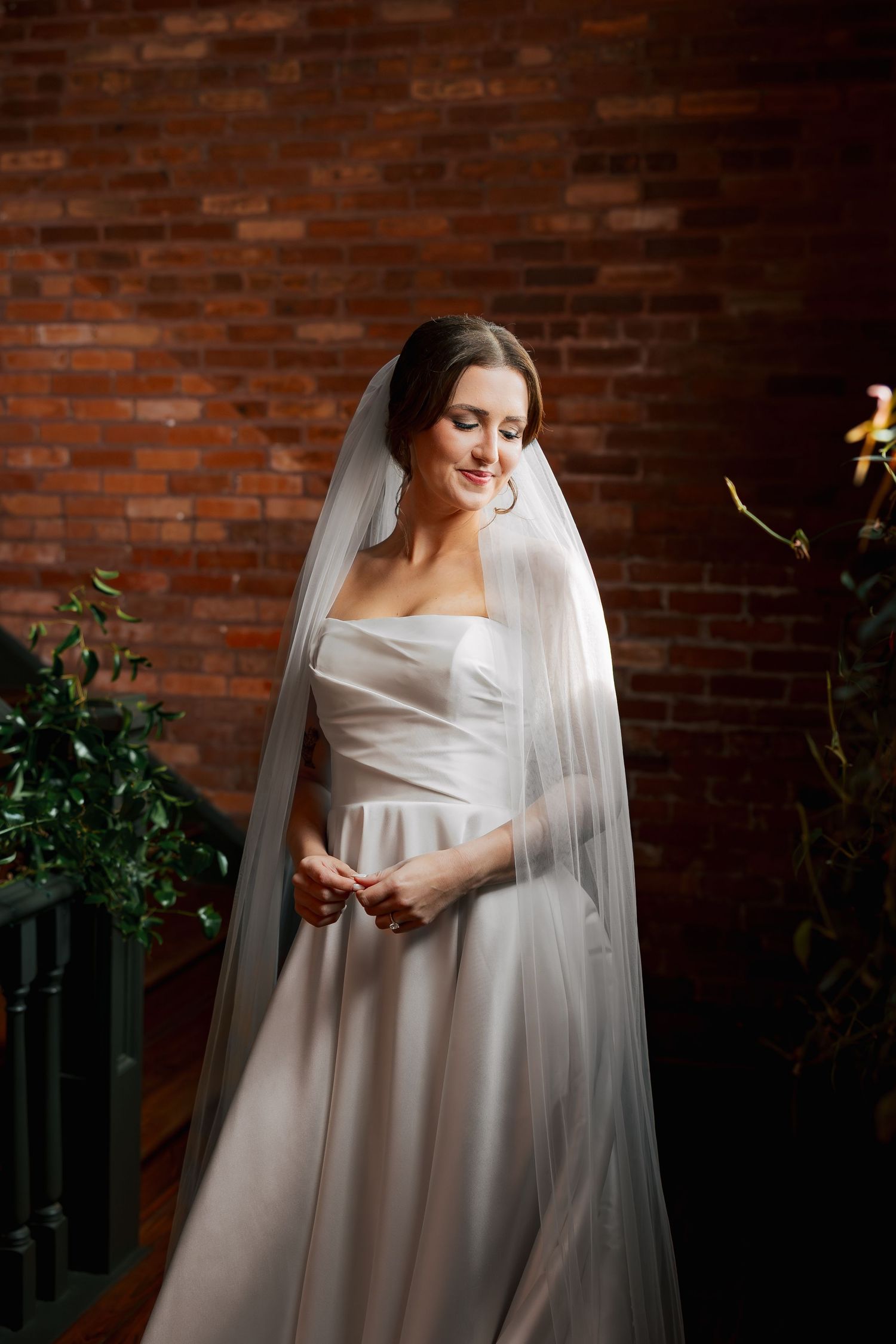 Elegant bride in strapless white gown and veil, profile view, smiling softly against gray backdrop.