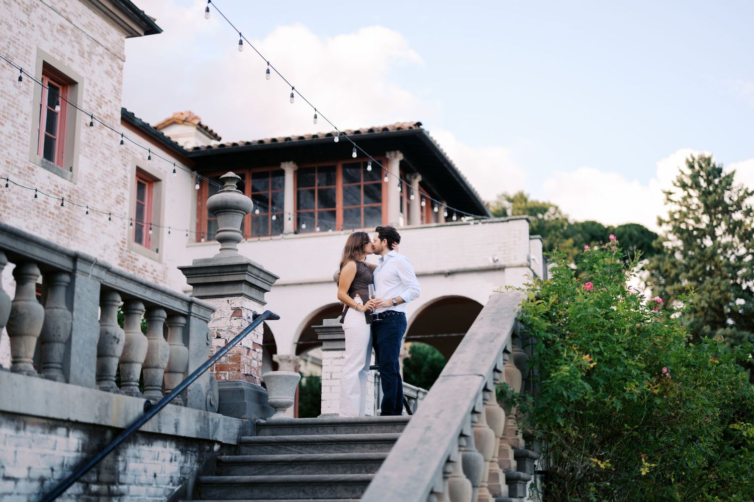 Couple sharing a romantic kiss on stone steps outside an elegant Spanish-style venue with string lights at dusk.