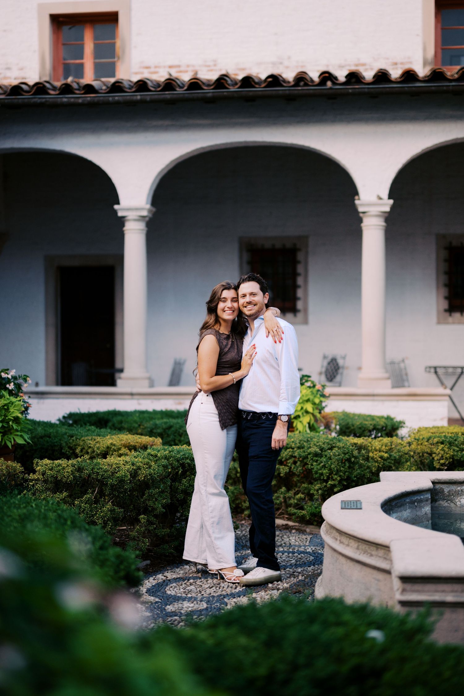 Couple embracing in elegant courtyard garden with arched colonnade and fountain at Mediterranean-style villa.