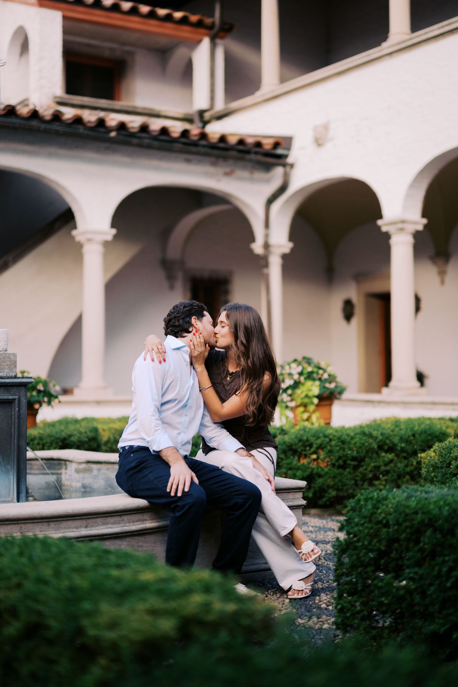 Couple sharing a romantic kiss while sitting on a stone bench in an elegant Italian courtyard with arched architecture.
