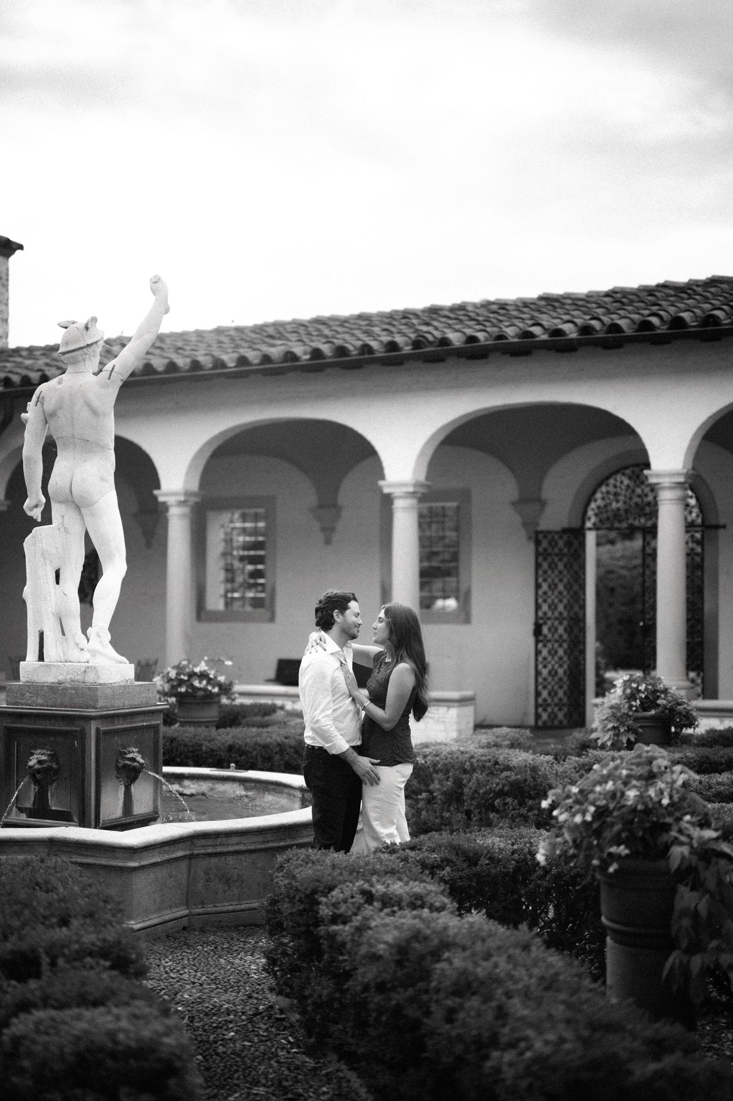 Black and white photo of a couple embracing in an Italian garden courtyard with a classical statue and arched building.