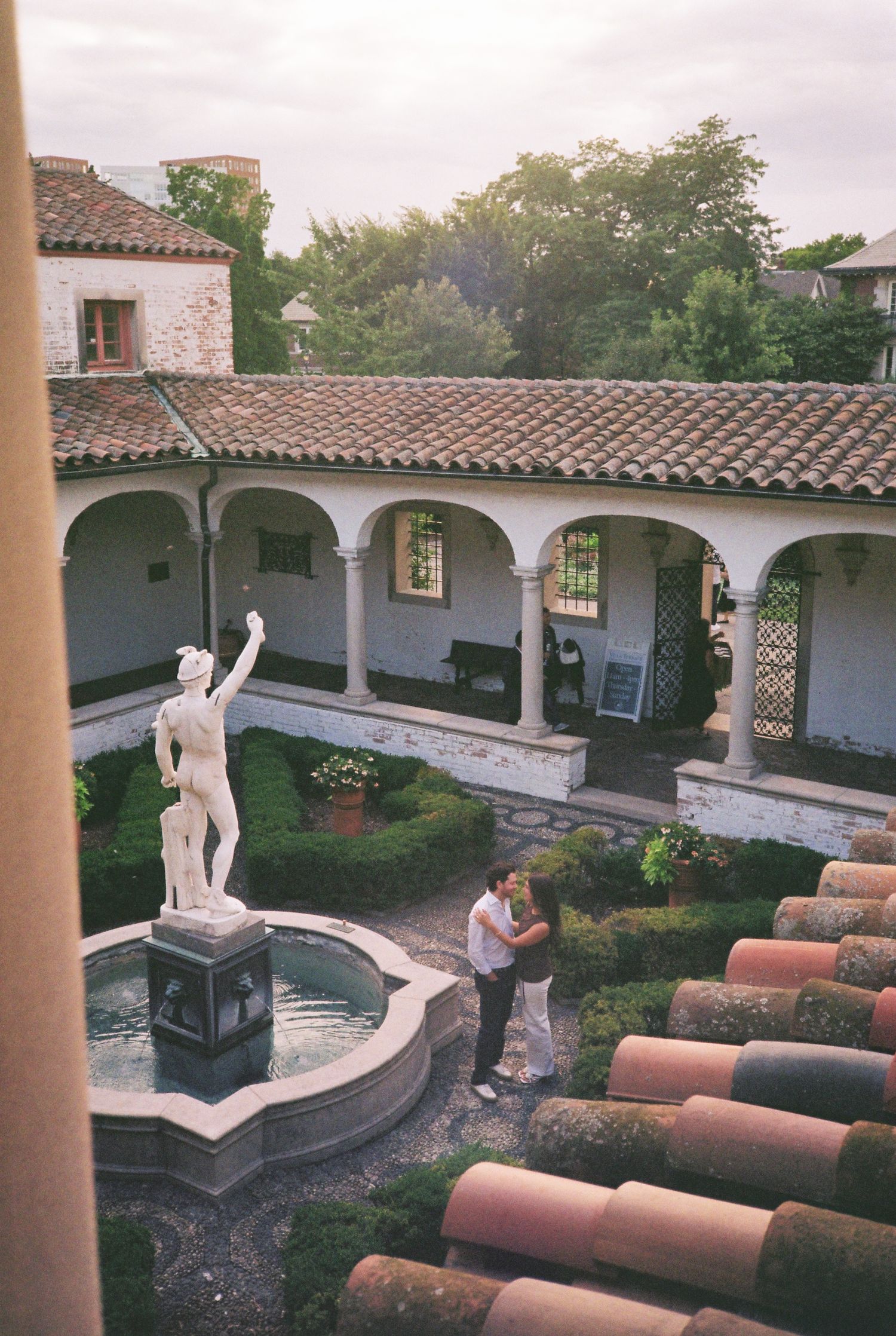 Couple embracing in a Mediterranean-style courtyard with a classical statue fountain, arched colonnade, and lush gardens.