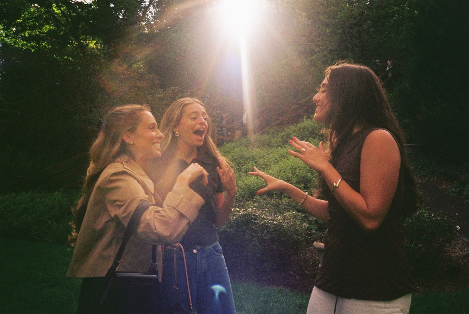 Three young women laughing together outdoors in sunlight, surrounded by lush green trees and garden.