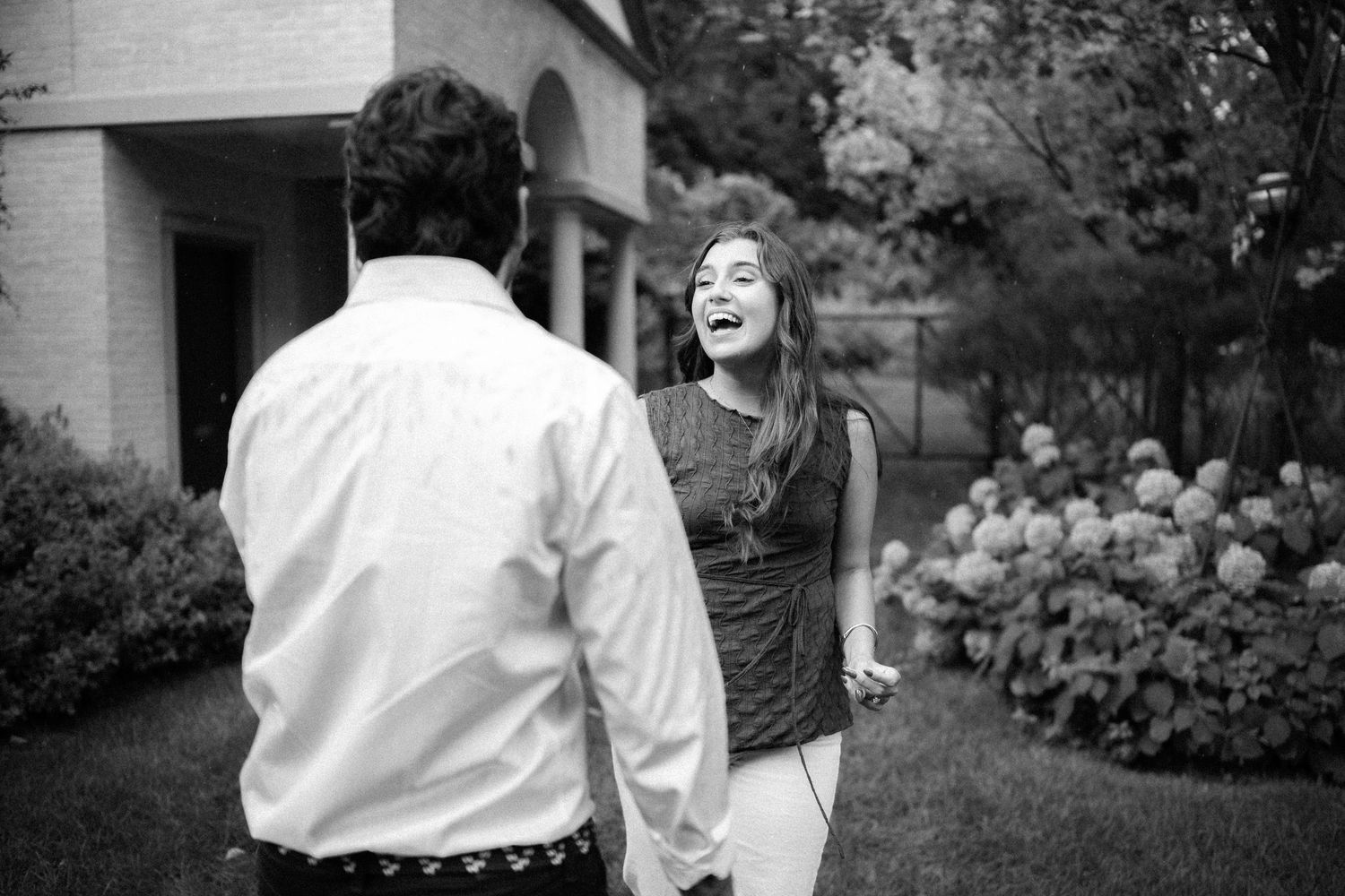 Black and white photo of a laughing couple holding hands in a garden outside a house.