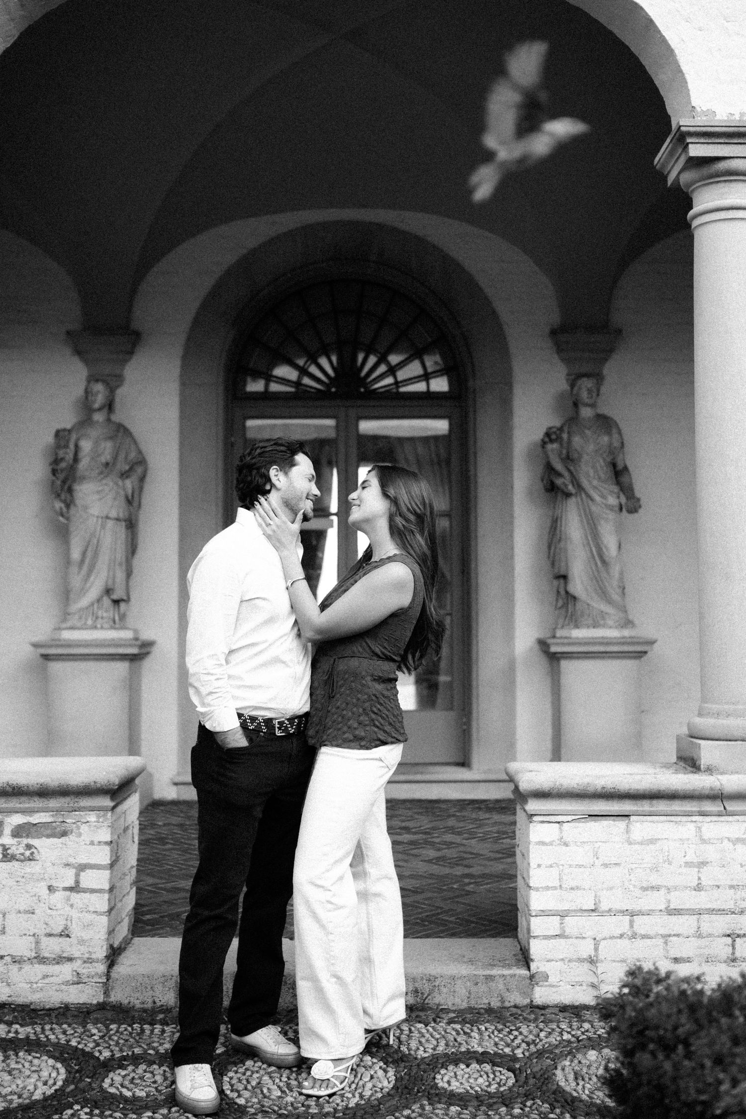 Black and white photo of a romantic couple embracing before an elegant arched building with classical statues and a dove in flight.