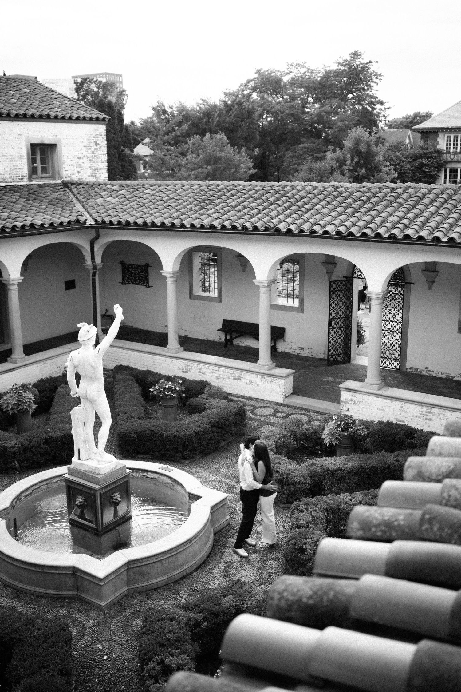 Black and white photo of a classical courtyard with arched colonnade, fountain statue, and two visitors exploring the garden.