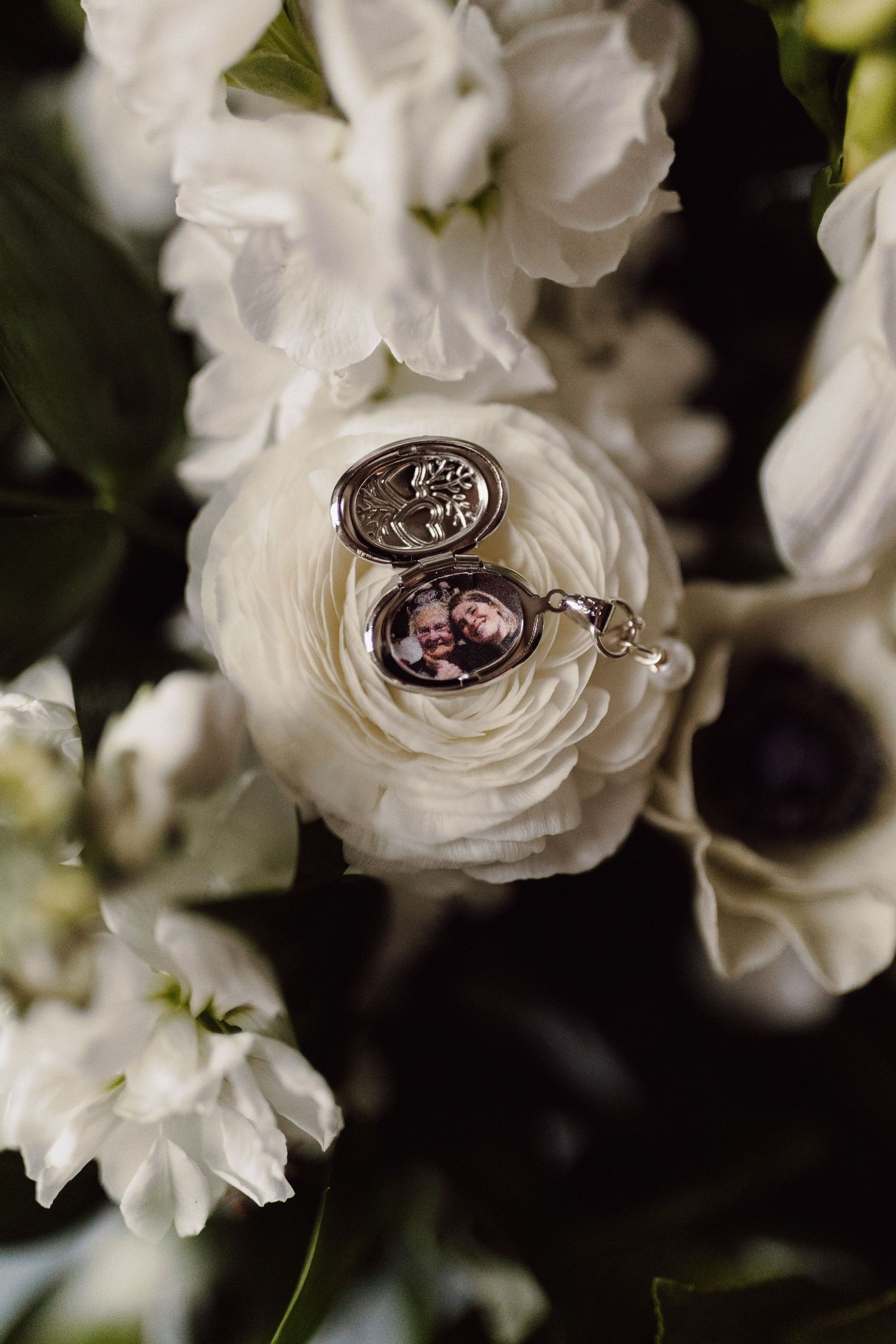 Open silver locket with couple photo resting in white ranunculus wedding bouquet.