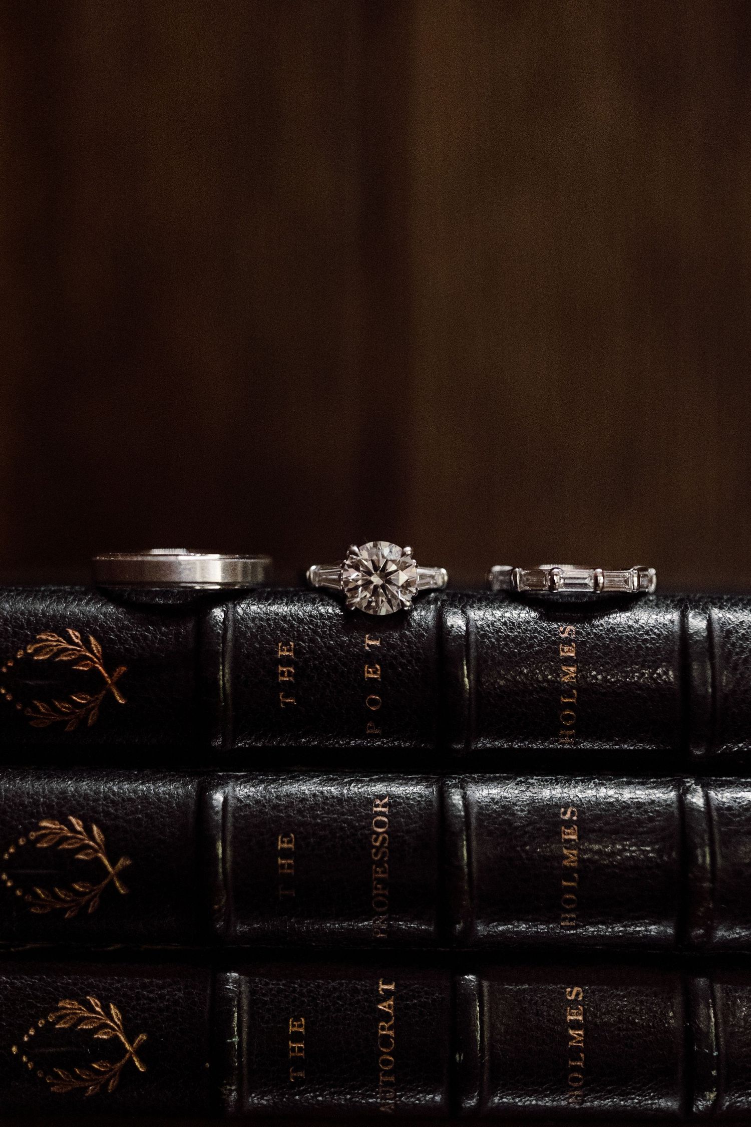 Three wedding rings displayed on antique leather-bound books with gold lettering in a dark, moody setting.