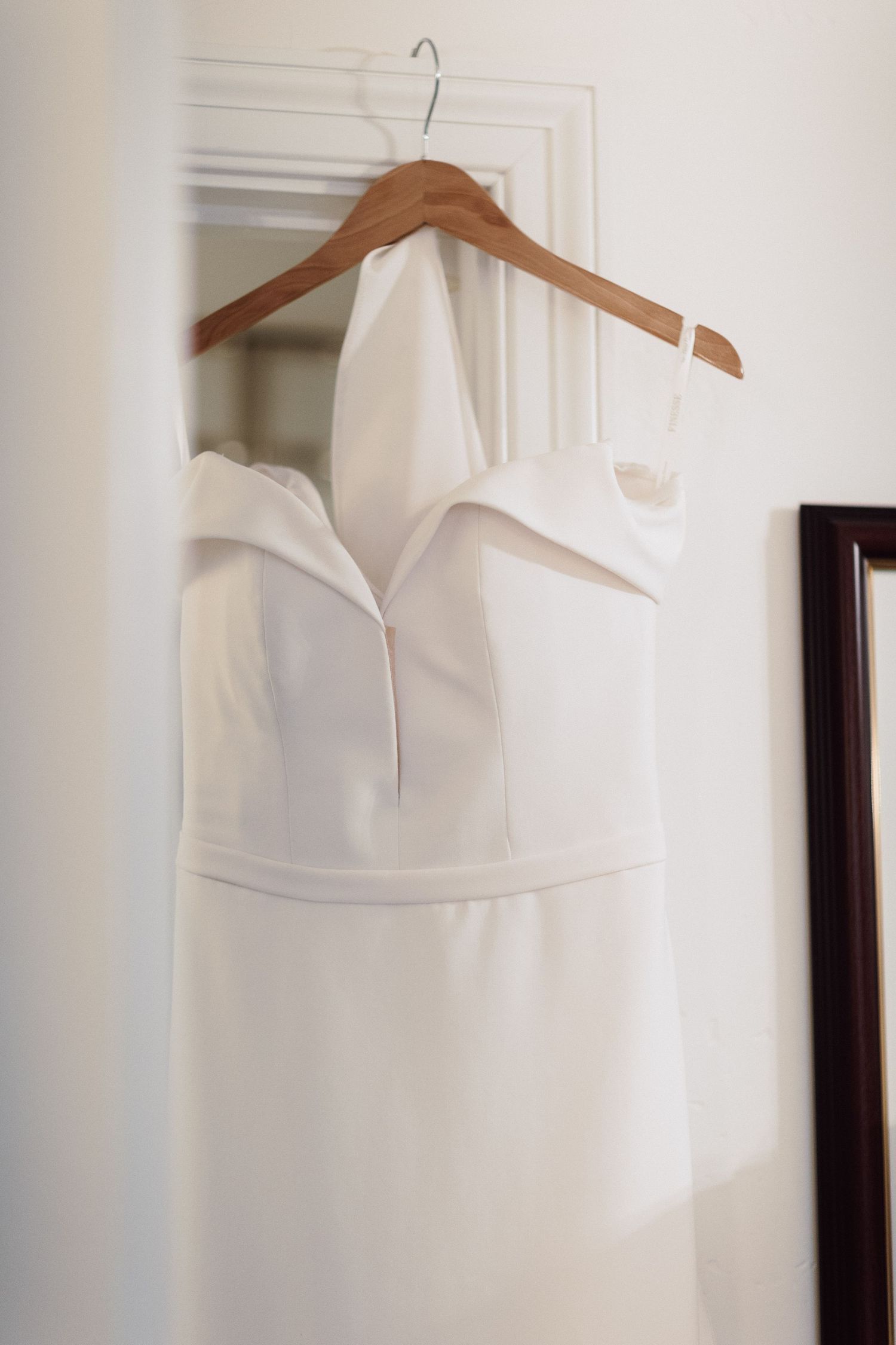 Elegant white halter neck wedding dress hanging on wooden hanger against white wall near mirror.