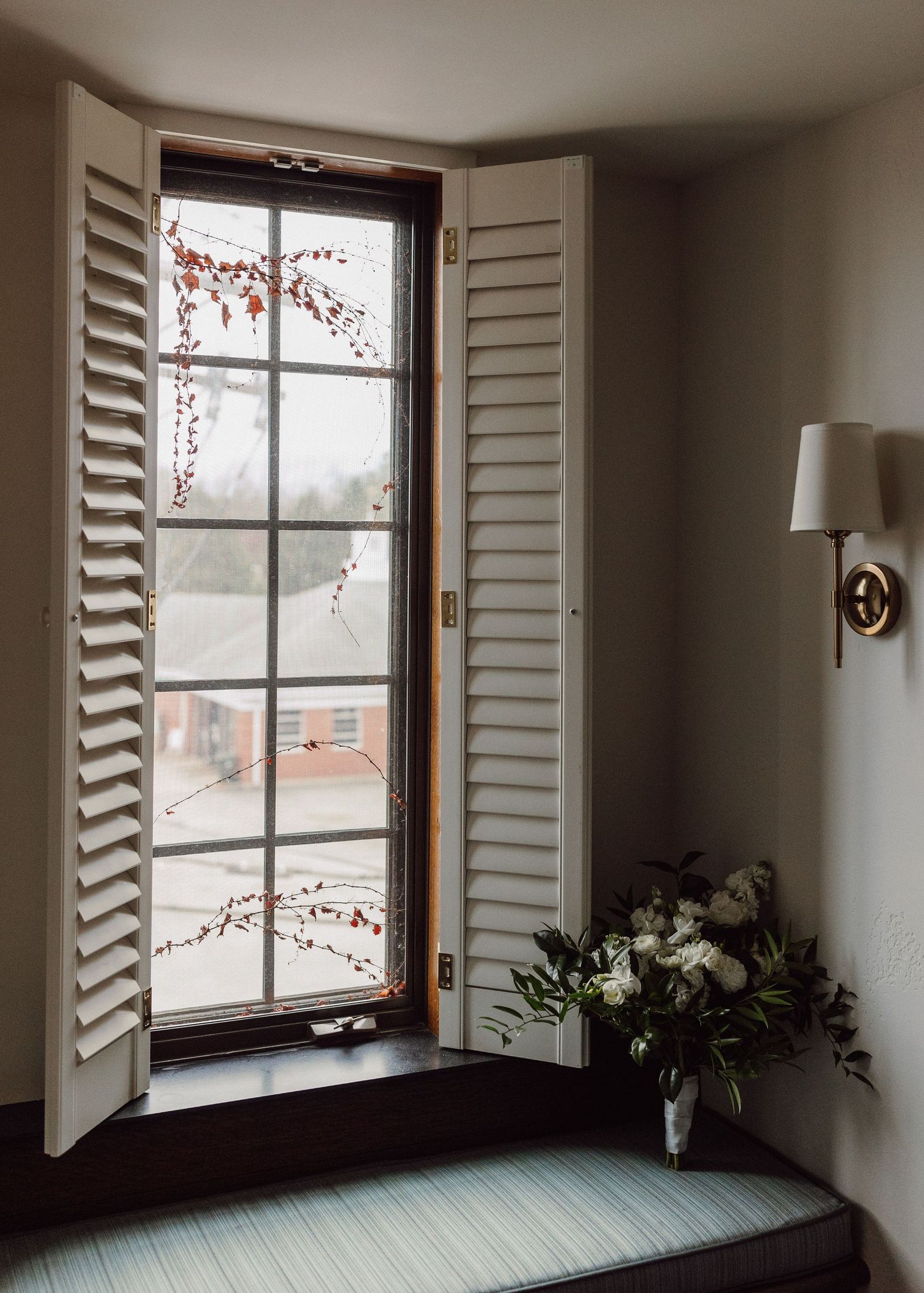 Elegant bay window with white plantation shutters, wall sconce, and white floral bouquet on window seat.