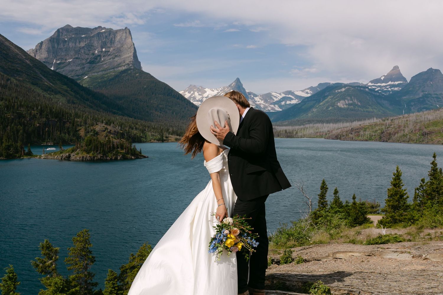 Couple sharing a kiss in wedding attire with a bouquet by a turquoise mountain lake under blue skies.