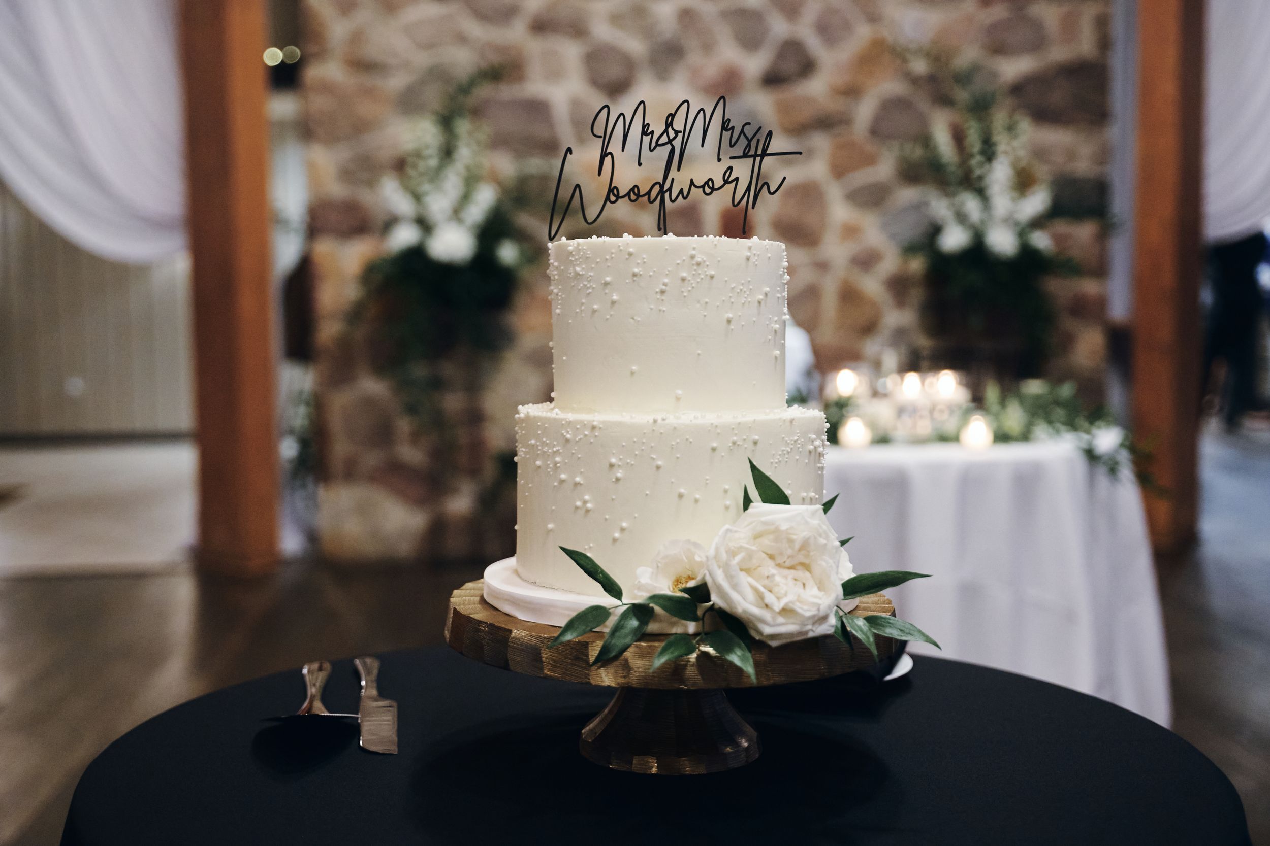 Elegant three-tier white wedding cake with floral topper reading Mr & Mrs Woodwork, decorated with roses on a gold stand.
