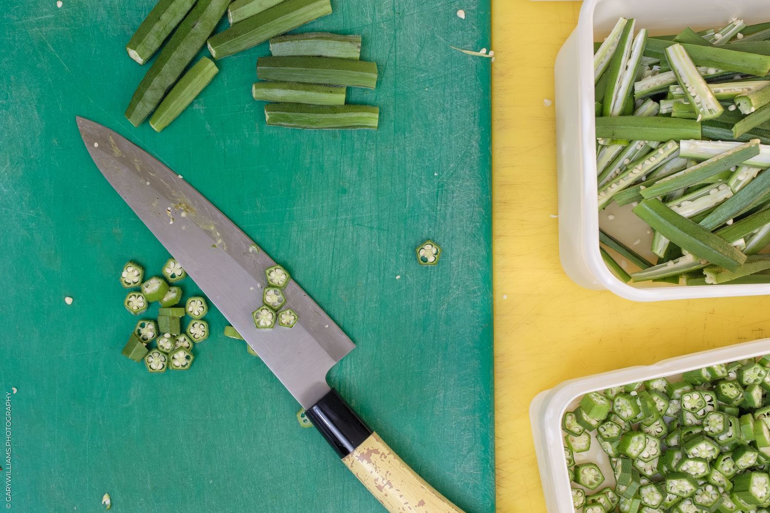 Fresh okra being sliced on a green cutting board with a chef knife, with cut pieces in white containers nearby.