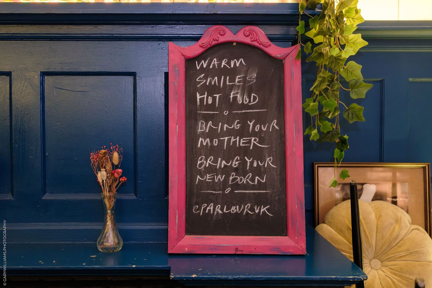 A pink-framed chalkboard sign with handwritten text on a blue wall in a cozy cafe or pub setting.