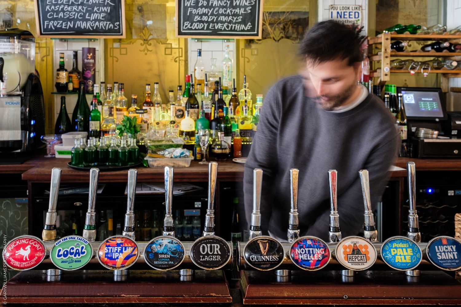 Bartender behind a pub bar with multiple beer taps including Guinness and Coors in a traditional British pub.