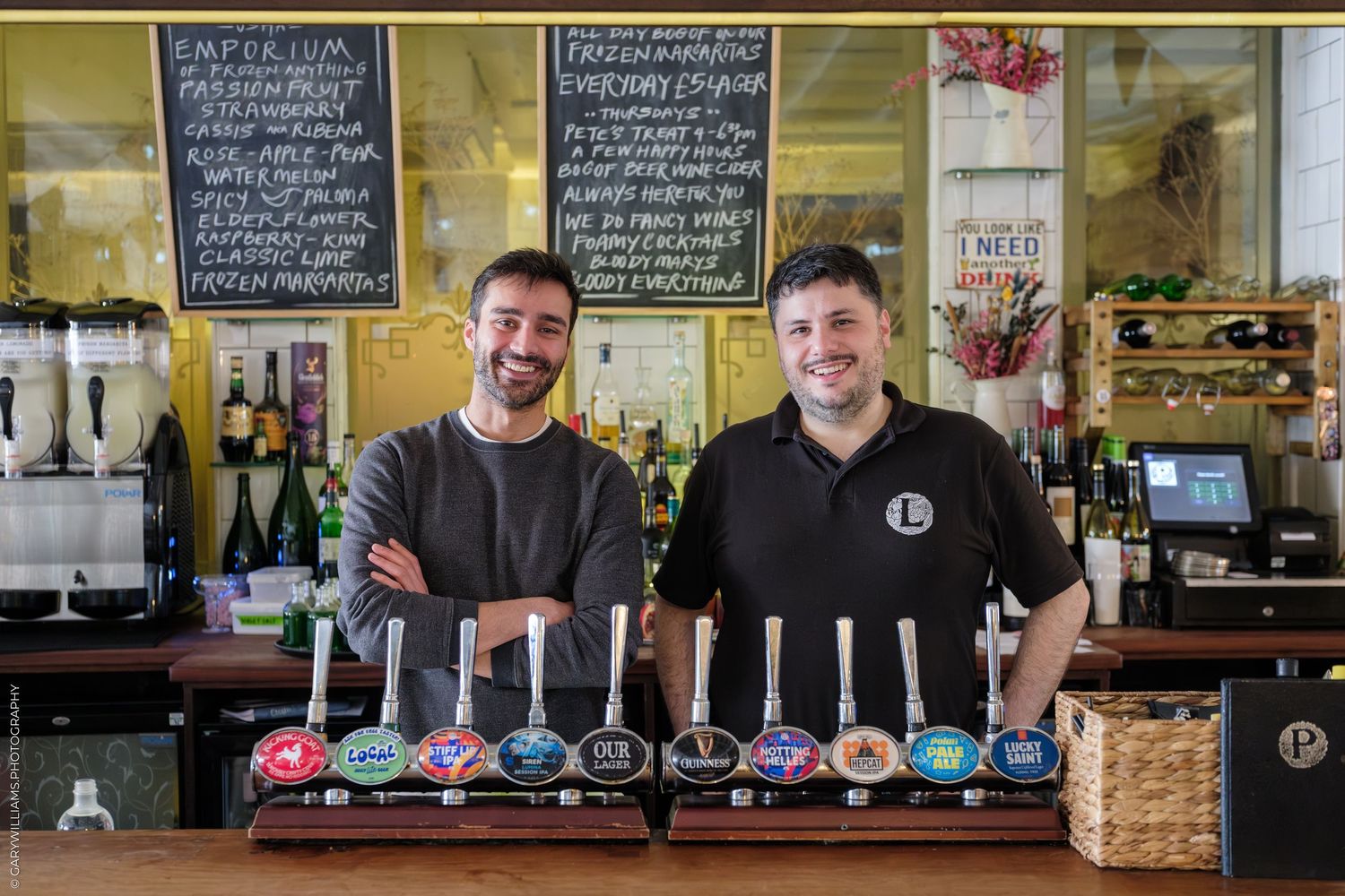 Two smiling bartenders stand behind a bar with colorful beer taps at a craft brewery pub.