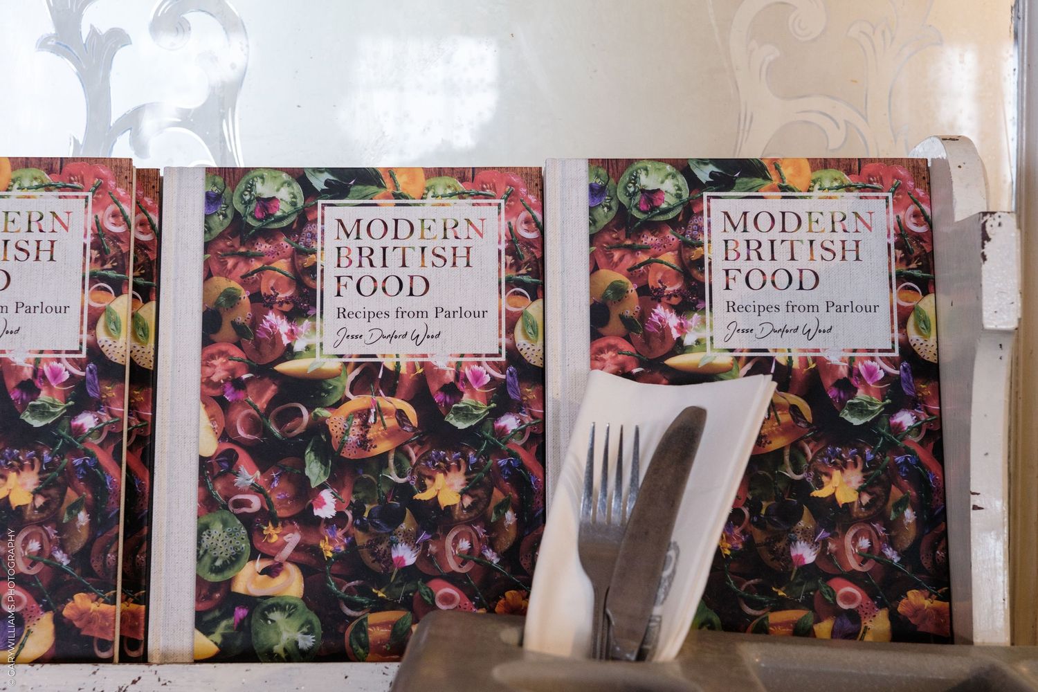 Three copies of Modern British Food cookbook displayed with a vintage fork and knife on a white surface.