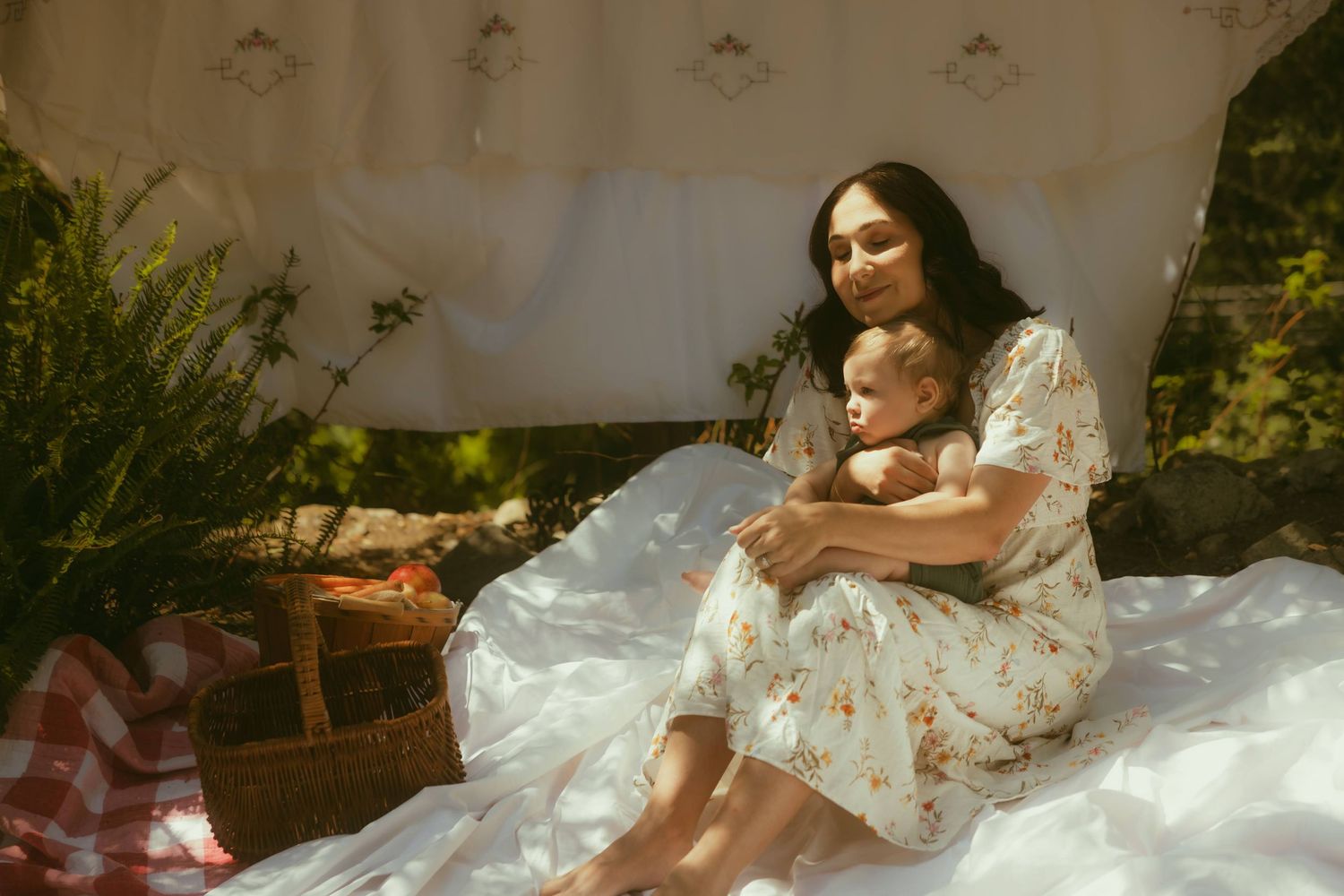 A mother in a floral dress holds her baby outdoors on a white blanket during a sunny picnic setting.