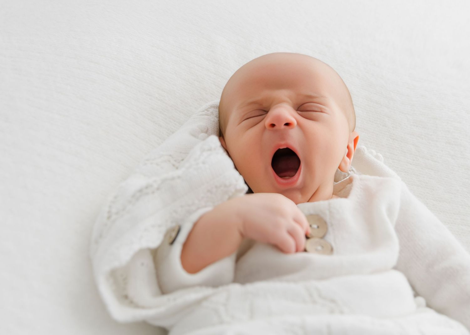 Newborn baby yawning while lying on white blanket, wearing white onesie, eyes closed, peaceful nursery photo.