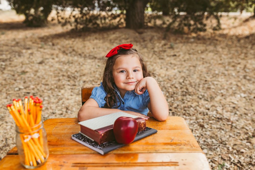 Young girl with red bow sits at wooden desk outdoors with apple, books, and pencils for back to school photos in yucaipa ca