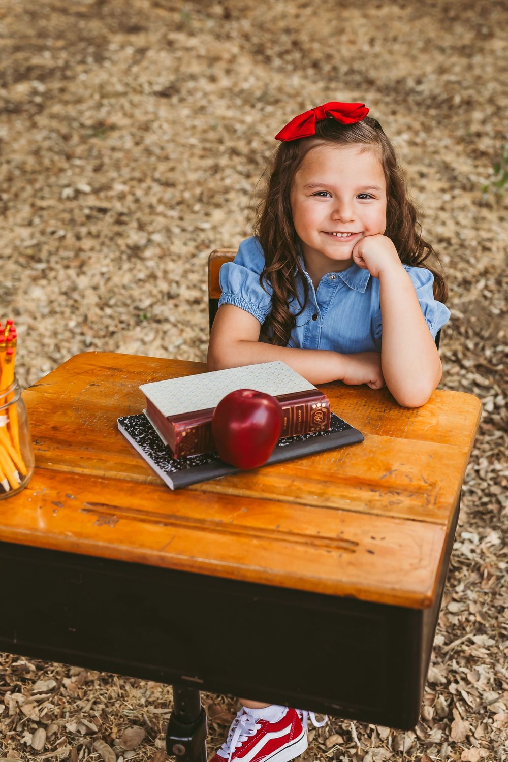 Smiling little girl with red hair bow poses at vintage school desk outside with apple and books with heidi grace photography