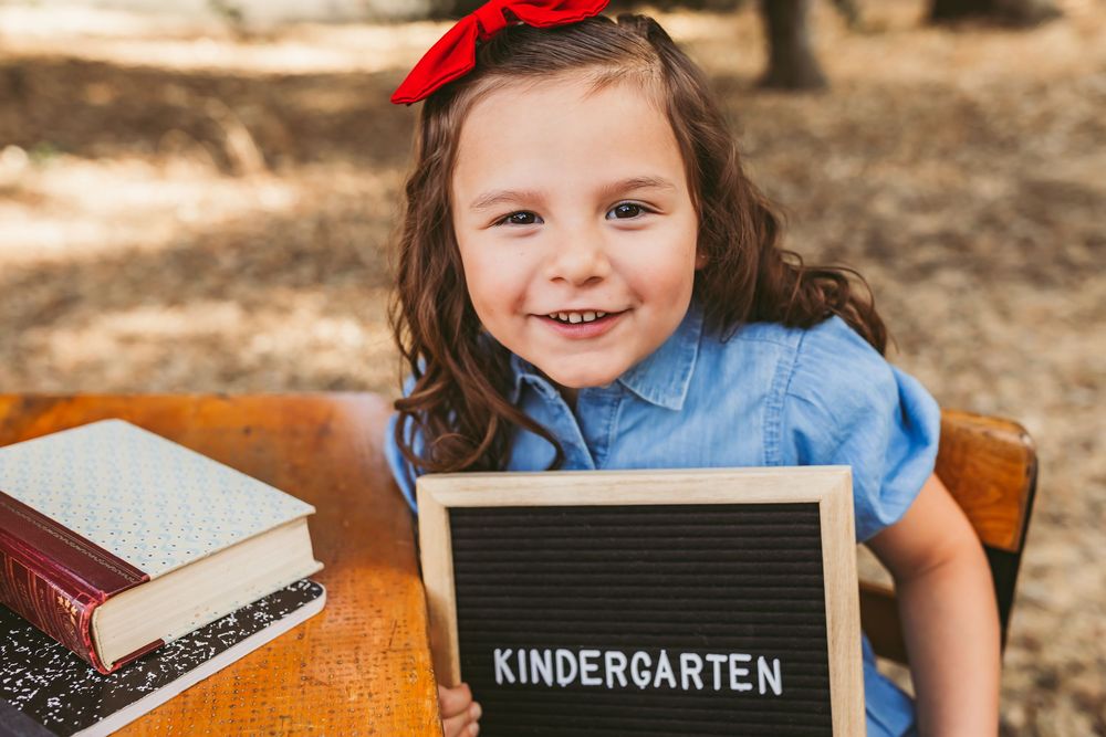 Happy young girl holds Kindergarten letter board sign at wooden desk outdoors for her back to school photos in yucaipa ca