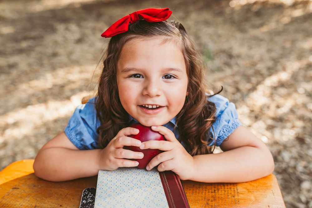 Cute little girl with red hair bow smiles while holding a red apple at a wooden desk outdoors.