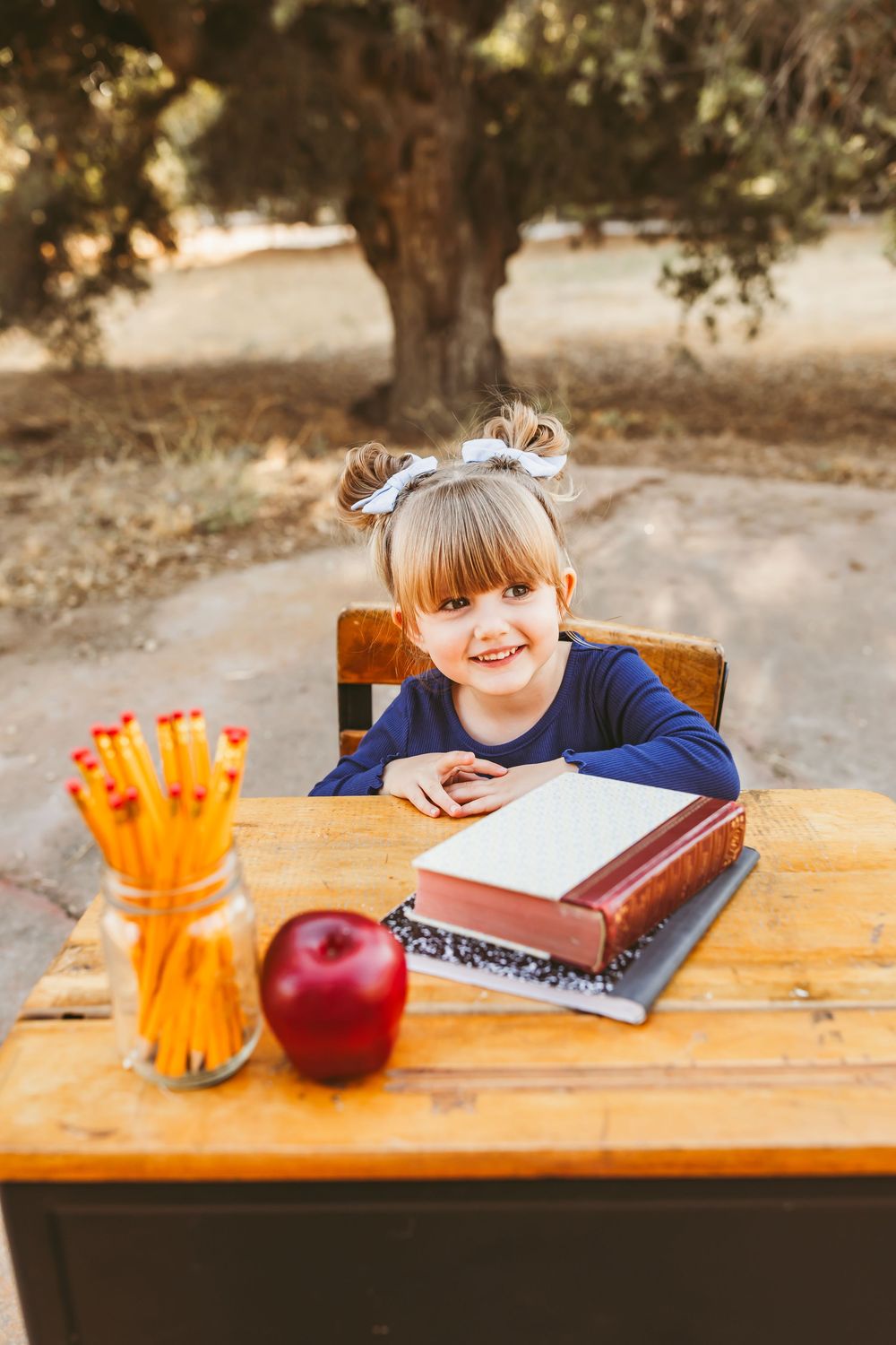 Smiling little girl sitting at a wooden desk outdoors with a book, red apple, and jar of pencils.