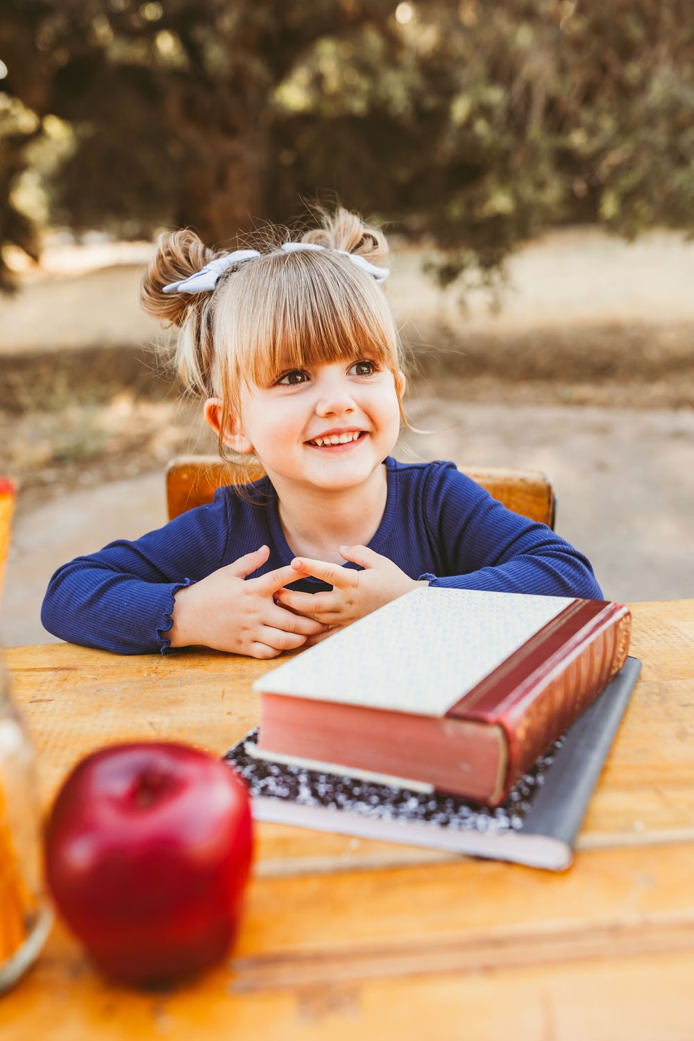 Smiling young girl sitting at outdoor wooden desk with red apple and books for her back to school mini session in yucaipa ca