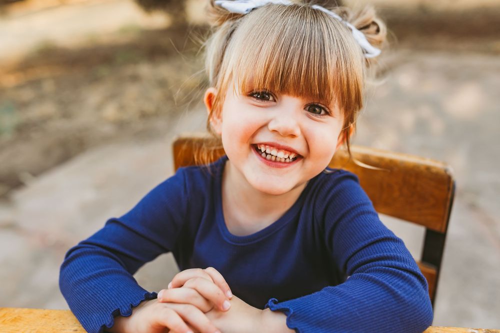 Smiling young girl with blonde hair and bow sitting outdoors in blue shirt.