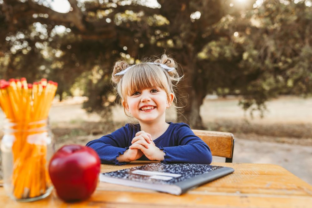 Happy little girl at outdoor desk with apple, pencils, and notebook ready for school photo shoot in the inland empire