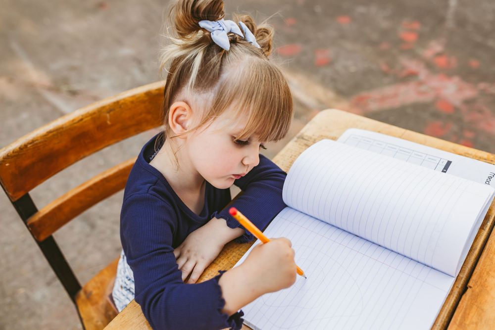 Young girl writing in notebook at wooden desk outdoors, focused on schoolwork.
