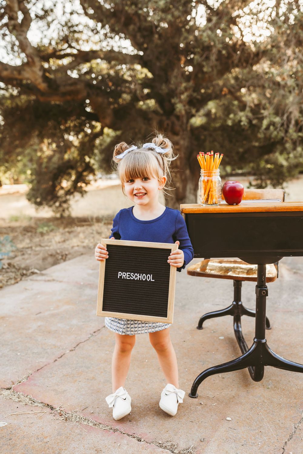 Smiling preschool girl holding a black letter board sign outdoors near a small desk with pencils and an apple.