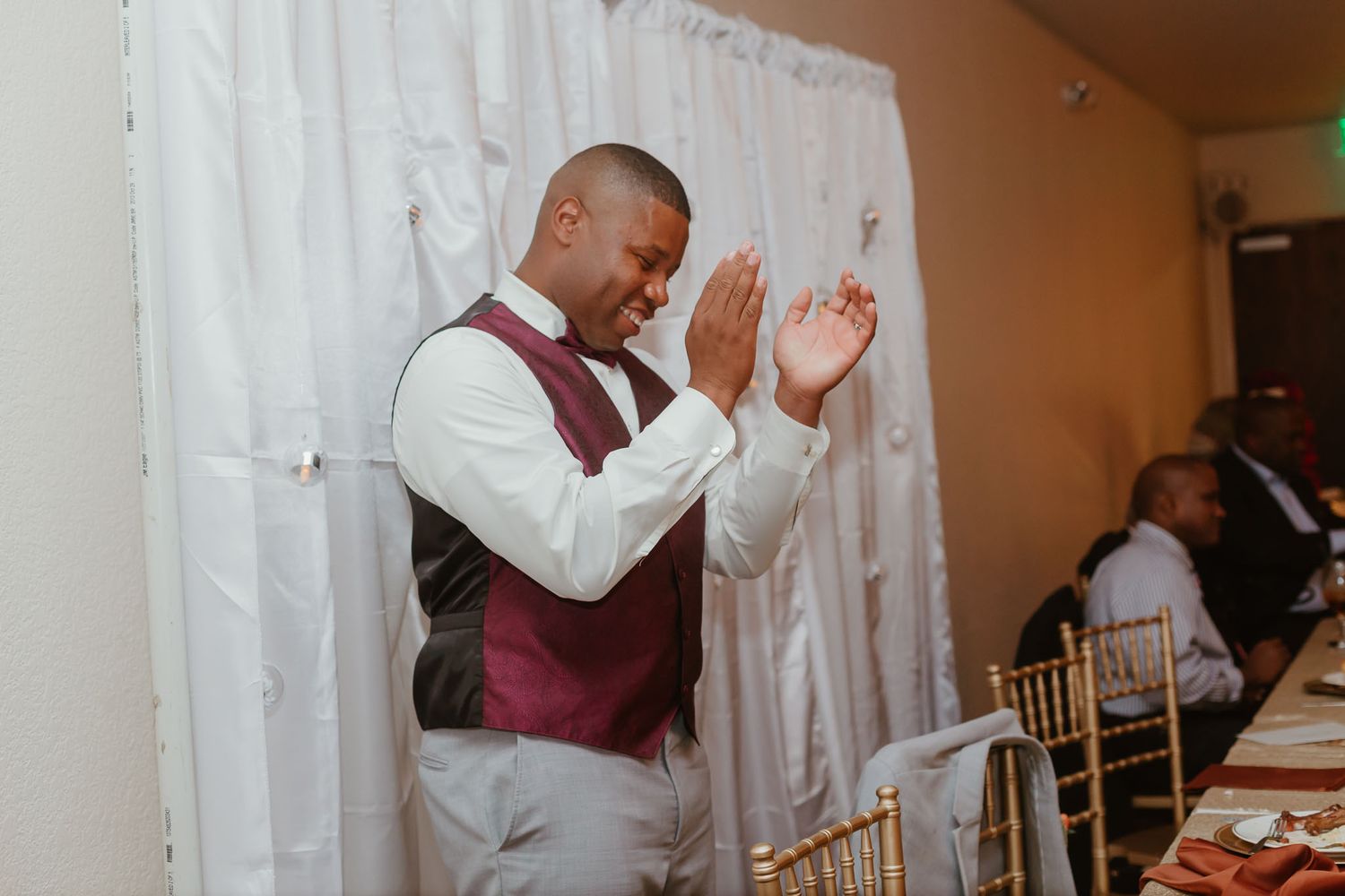 A man in a maroon vest and white shirt clapping his hands at an elegant wedding reception.
