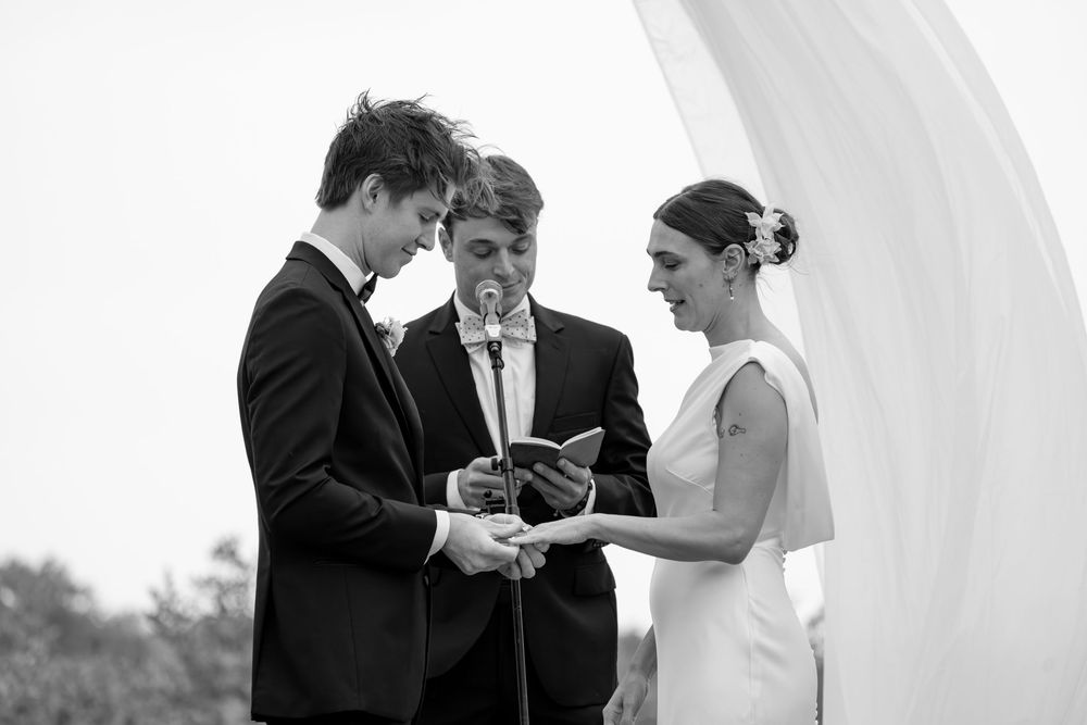Black and white wedding ceremony photo of bride and groom exchanging vows with officiant outdoors.