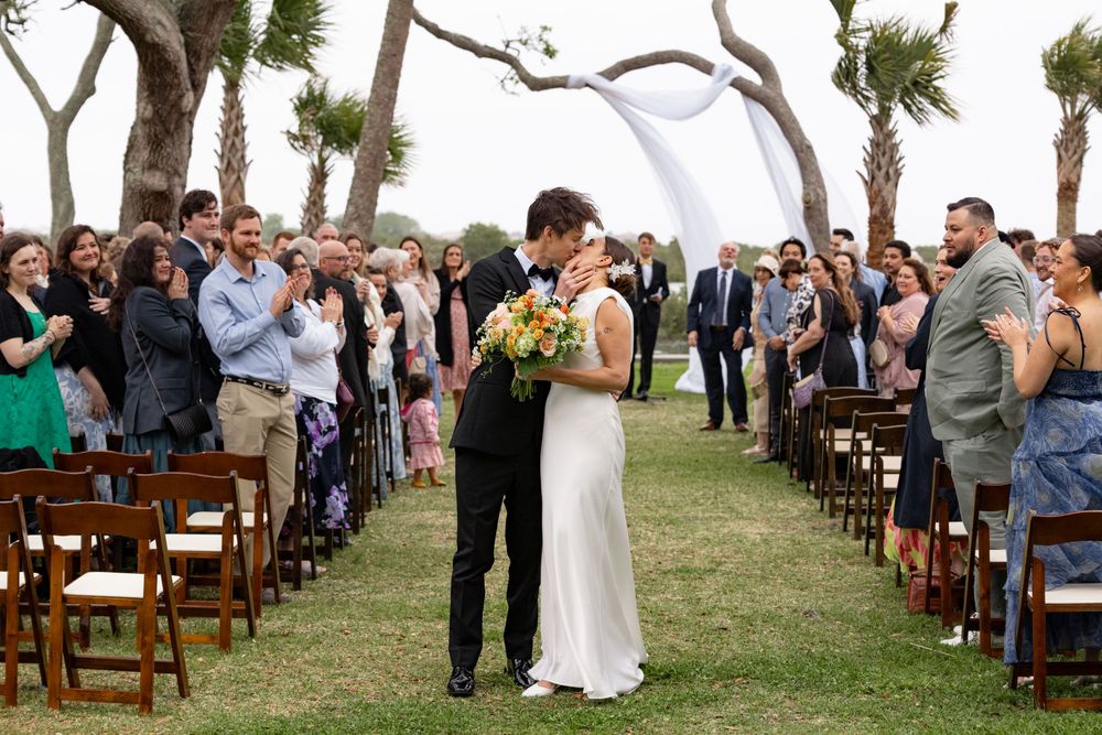 Bride and groom share first kiss at outdoor wedding ceremony with guests applauding along decorated aisle.