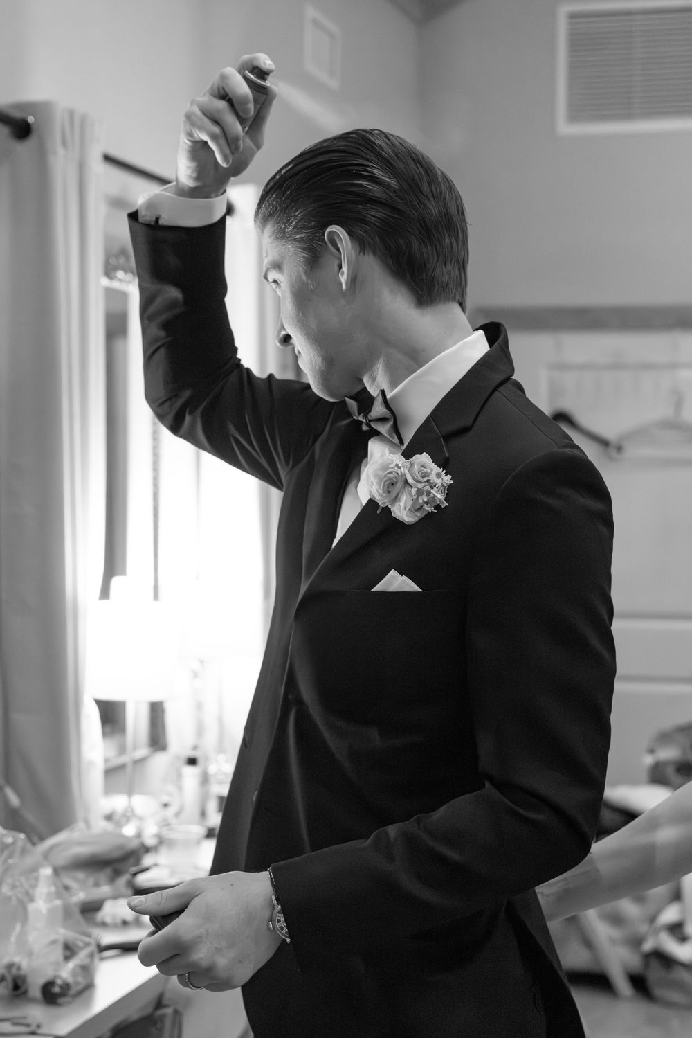 Groom in black tuxedo with boutonniere adjusting hair in black and white wedding preparation photo.