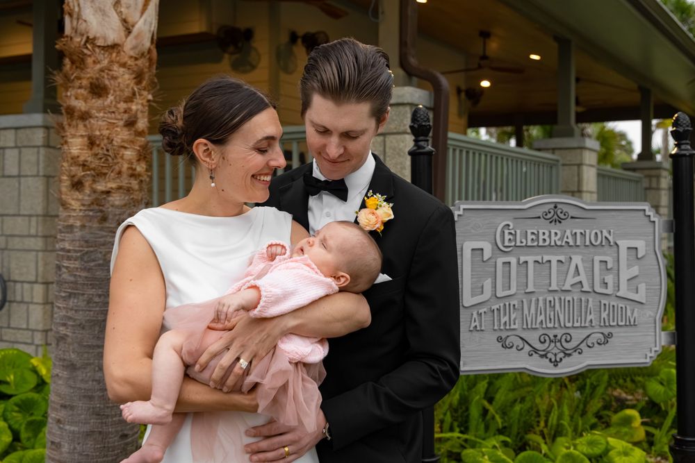 Newlywed couple in wedding attire tenderly holding their newborn baby outside Celebration Cottage.