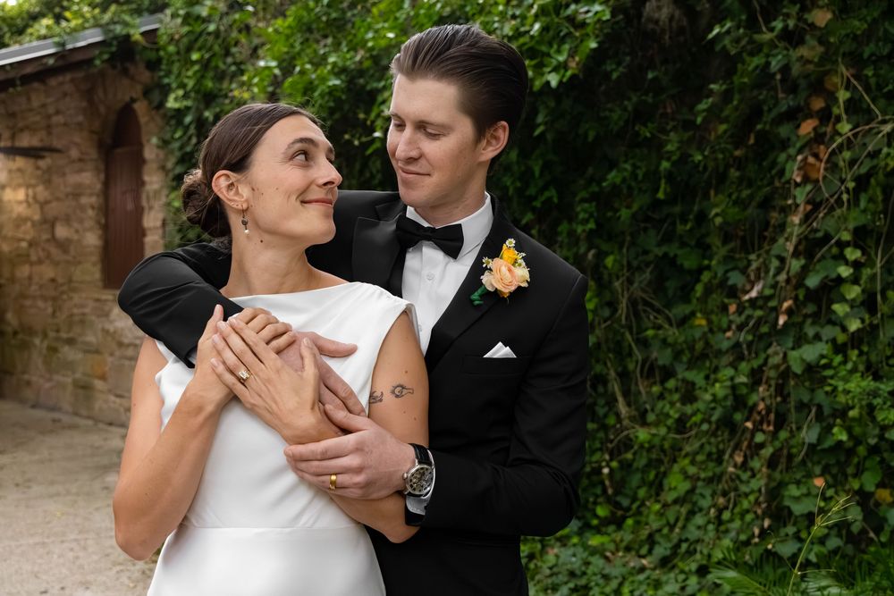 A bride and groom embrace outdoors, smiling at each other, groom in black tuxedo with yellow boutonniere.