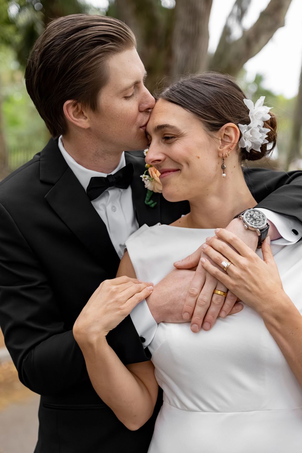 Groom in black tuxedo kissing bride on cheek as she smiles, both wearing wedding rings outdoors.