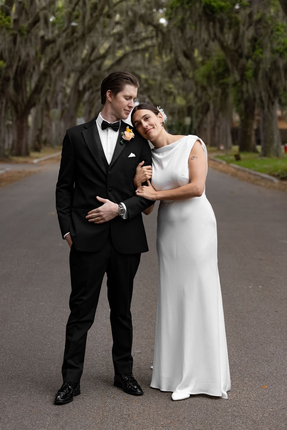A couple in wedding attire poses on a tree-lined road, the bride in white and groom in a black tuxedo.