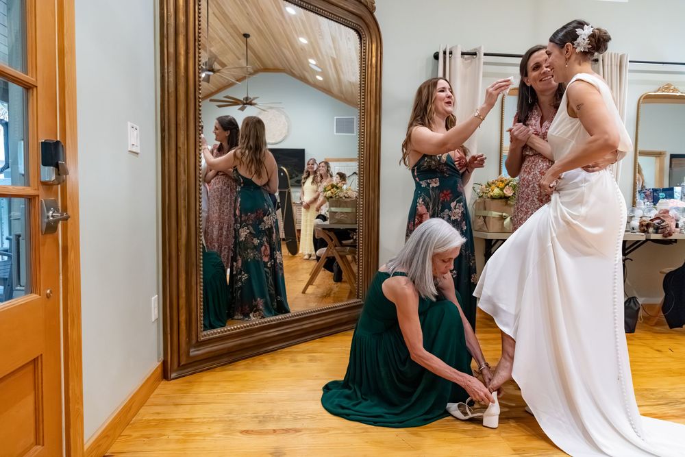Bride getting ready with bridesmaids helping with dress and shoes in a room with a large mirror.
