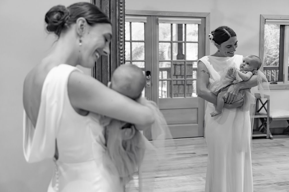 Black and white photo of two brides in white gowns holding babies at a wedding venue.