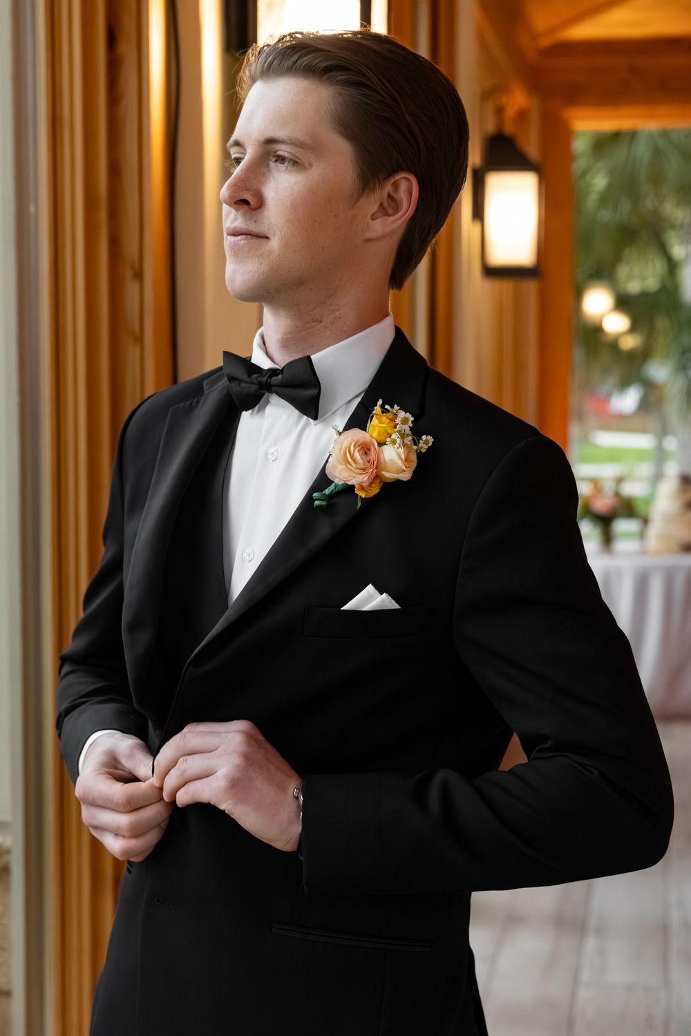 A dapper groom in a black tuxedo with bow tie and floral boutonniere buttons his jacket while gazing thoughtfully.