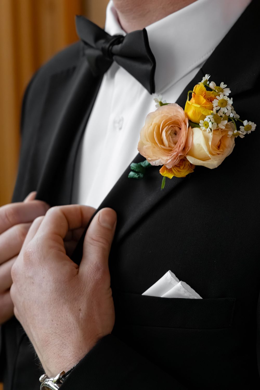 Man in black tuxedo with bow tie adjusting boutonniere of peach roses and yellow flowers on lapel with white pocket square.